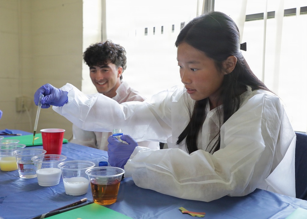 Two young students wearing white lab coats and purple nitrile gloves are engaged in a hands-on science activity at a table covered with a blue cloth. The foreground student, a girl with long dark hair, is using a dropper and holding a small vial while working with various liquids in clear plastic cups. A male student in the background is smiling and observing the activity.