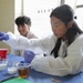 Two young students wearing white lab coats and purple nitrile gloves are engaged in a hands-on science activity at a table covered with a blue cloth. The foreground student, a girl with long dark hair, is using a dropper and holding a small vial while working with various liquids in clear plastic cups. A male student in the background is smiling and observing the activity.