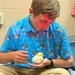 A young male student sits cross-legged on a carpeted floor holding a small yellow baby chick in his cupped hands. He is wearing a bright blue polo shirt with a rubber duck surfing pattern and appears to be in a school or institutional setting. A red heat lamp is casting light on his face and head, suggesting the chick requires warmth.