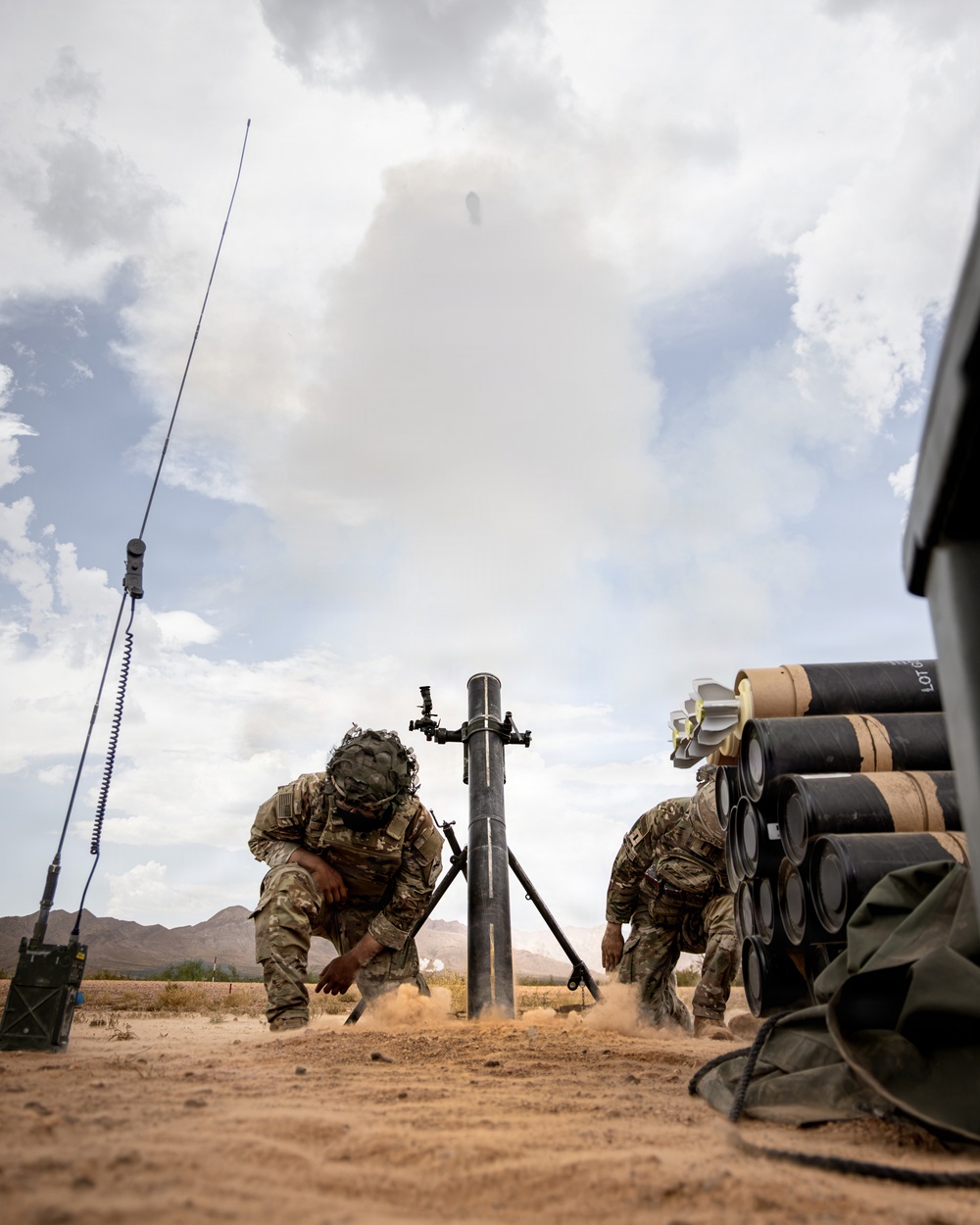 Two soldiers in camouflage uniforms are operating a mortar system in a desert training environment. A round is visibly in flight above the mortar tube immediately after firing, with smoke and dust displacement evident. Multiple stacked mortar rounds are visible to the right, along with a radio antenna assembly to the left.