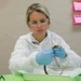 A young woman in a laboratory or classroom setting is performing a dissection, wearing full protective gear including a white lab coat, blue nitrile gloves, and clear safety glasses. She is focused on handling what appears to be a specimen (likely a small animal such as a frog or similar organism) over a metal dissection tray. The setting appears to be an educational environment, suggested by colorful papers posted on the wall in the background.