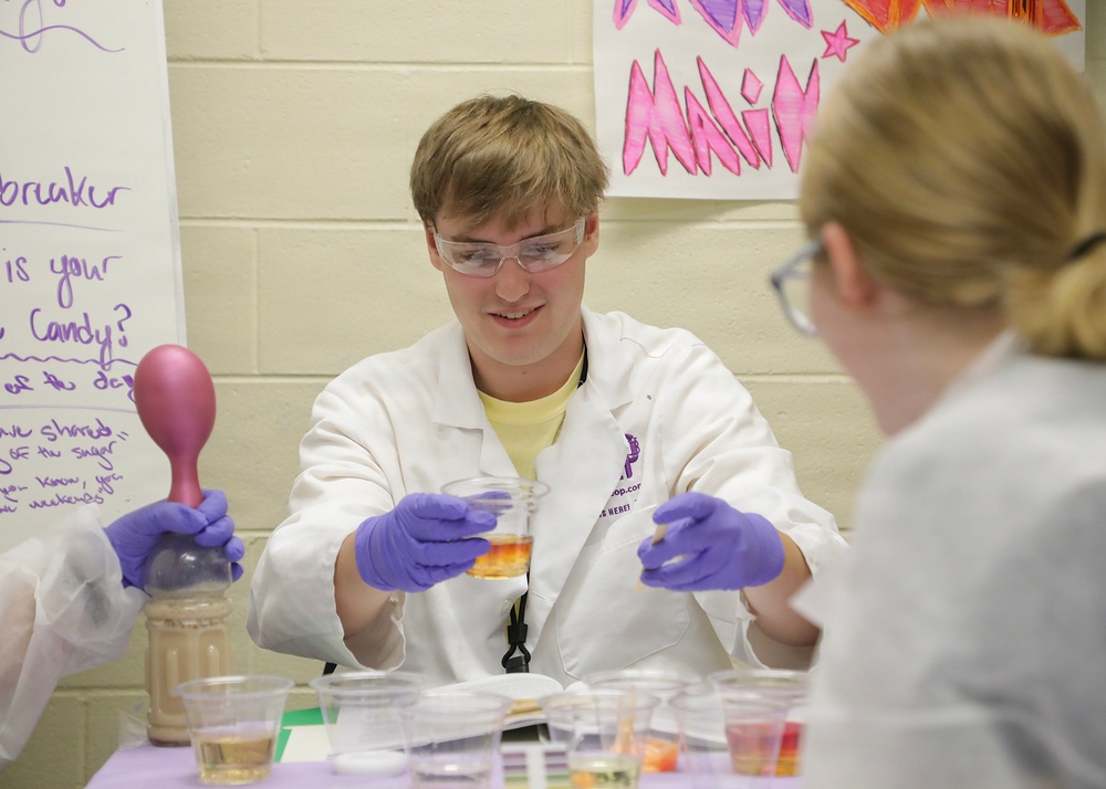 A young male in a white lab coat, safety goggles, and purple nitrile gloves is conducting a science demonstration or experiment at a table. He appears to be presenting materials to another person seated across from him, likely in an educational or outreach setting. The environment suggests a school or community science event.