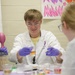 A young male in a white lab coat, safety goggles, and purple nitrile gloves is conducting a science demonstration or experiment at a table. He appears to be presenting materials to another person seated across from him, likely in an educational or outreach setting. The environment suggests a school or community science event.