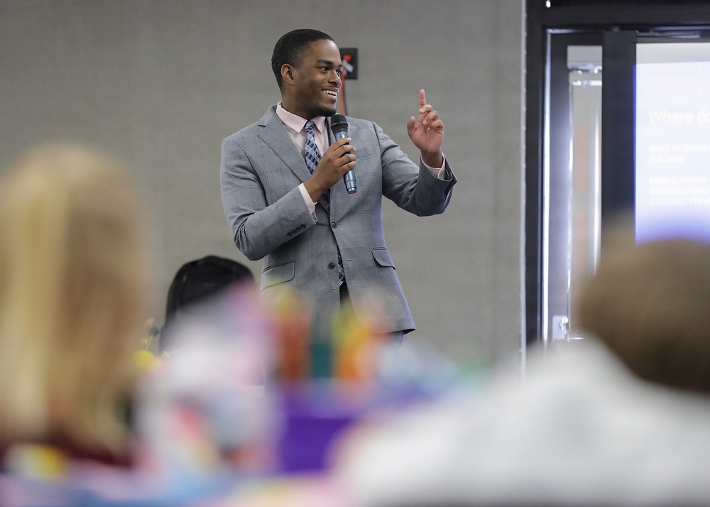 A young man in a gray suit is delivering a speech or presentation to an audience in what appears to be a conference or event hall. He is holding a microphone in one hand and raising his index finger with the other, smiling confidently. A projection screen is partially visible in the background to the right.