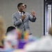 A young man in a gray suit is delivering a speech or presentation to an audience in what appears to be a conference or event hall. He is holding a microphone in one hand and raising his index finger with the other, smiling confidently. A projection screen is partially visible in the background to the right.