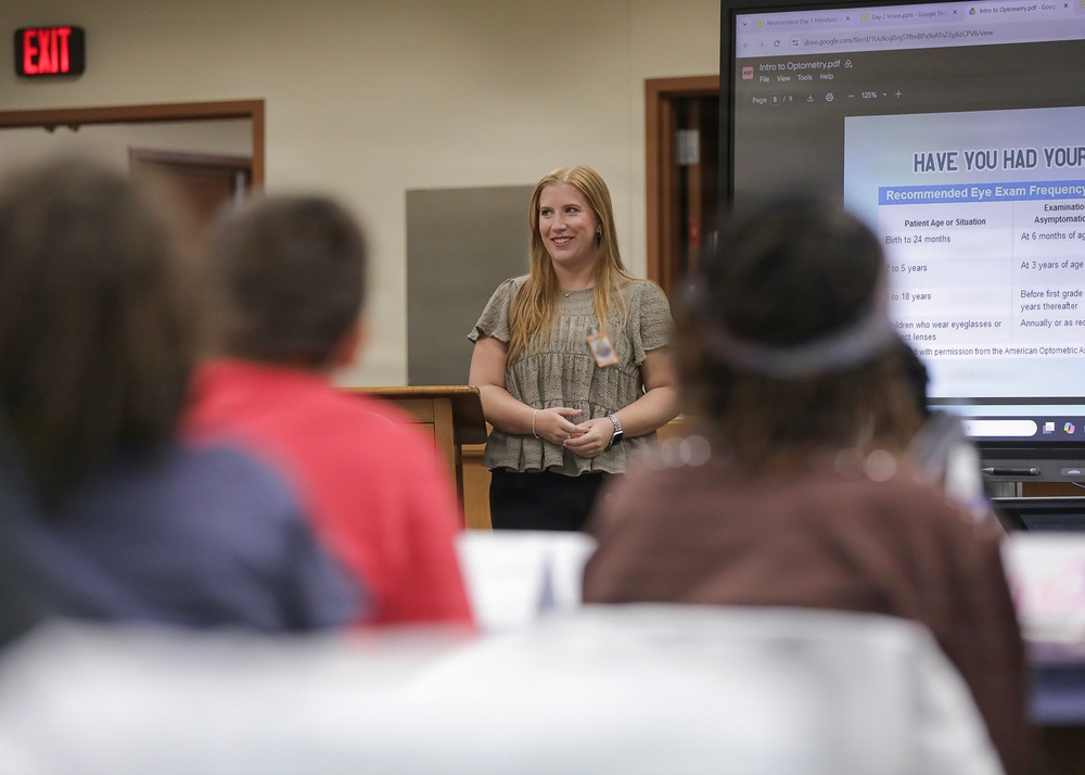 A woman with long blonde hair is presenting at the front of a classroom or meeting room, standing near a podium. Behind her, a large monitor displays a PDF titled 'Intro to Optometry' showing a slide about recommended eye exam frequency. An audience of several people is seated at tables facing the presenter.