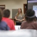 A woman with long blonde hair is presenting at the front of a classroom or meeting room, standing near a podium. Behind her, a large monitor displays a PDF titled 'Intro to Optometry' showing a slide about recommended eye exam frequency. An audience of several people is seated at tables facing the presenter.