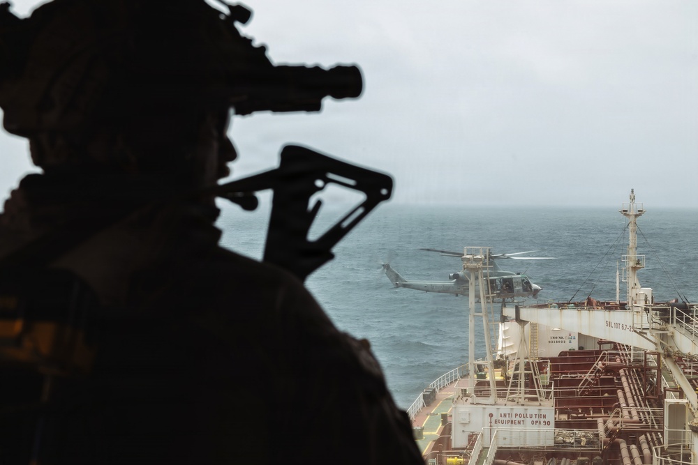 A silhouetted armed military operator wearing a helmet with night-vision device mounts and carrying a weapon observes a helicopter hovering near a large commercial tanker vessel at sea. The scene appears to depict a maritime interdiction or visit, board, search, and seizure (VBSS) operation. Overcast conditions and open ocean are visible in the background.