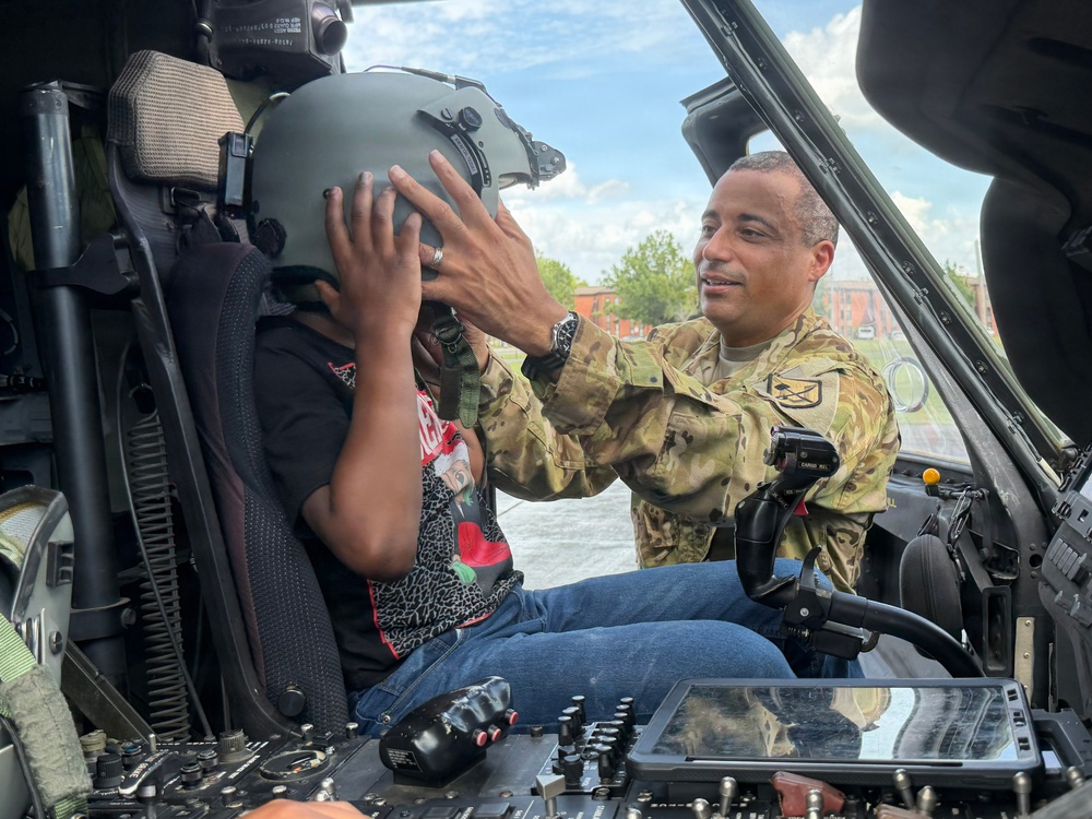 A military service member in OCP (Operational Camouflage Pattern) uniform is placing a flight helmet on a civilian child seated in the cockpit of a military helicopter. The interaction appears to be part of a public outreach or community engagement event, allowing civilians to experience the aircraft up close. The setting appears to be an outdoor static display or air show.