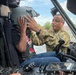 A military service member in OCP (Operational Camouflage Pattern) uniform is placing a flight helmet on a civilian child seated in the cockpit of a military helicopter. The interaction appears to be part of a public outreach or community engagement event, allowing civilians to experience the aircraft up close. The setting appears to be an outdoor static display or air show.