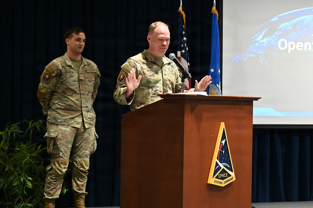 Two military personnel in U.S. Space Force/Army camouflage uniforms are at a formal event. The primary speaker stands at a wooden podium bearing the Space Systems Command insignia, gesturing with both hands while addressing an audience. A projection screen behind them displays partial text reading 'Open...' alongside an image of Earth.