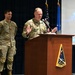 Two military personnel in U.S. Space Force/Army camouflage uniforms are at a formal event. The primary speaker stands at a wooden podium bearing the Space Systems Command insignia, gesturing with both hands while addressing an audience. A projection screen behind them displays partial text reading 'Open...' alongside an image of Earth.