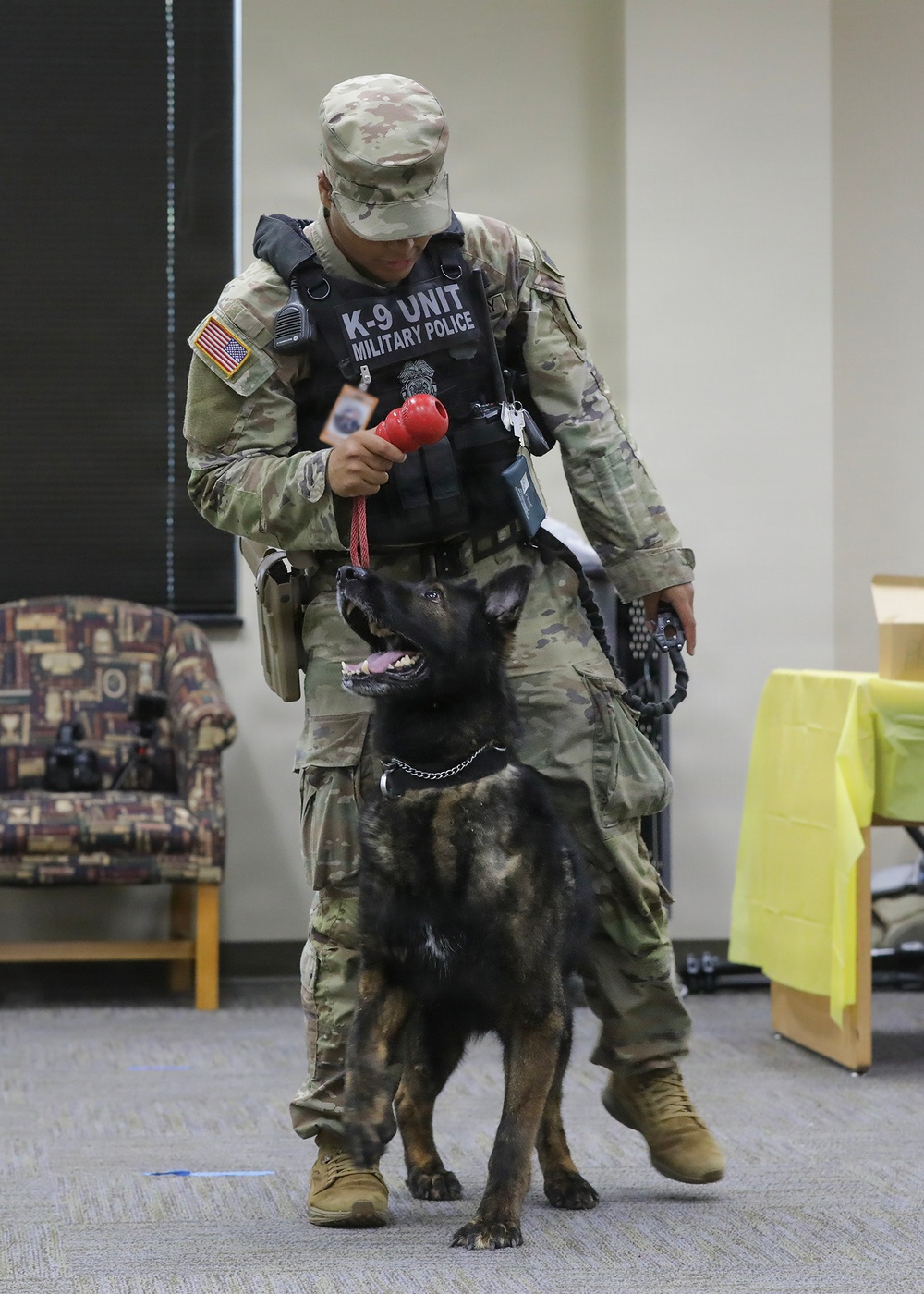 A U.S. Army Military Police K-9 handler in full camouflage uniform and tactical vest is conducting a training or demonstration session indoors with a dark-coated Belgian Malinois or German Shepherd. The handler is holding a red Kong toy as a reward tool while the dog looks up attentively. The setting appears to be an indoor facility such as a community center or meeting room.