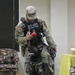 A U.S. Army Military Police K-9 handler in full camouflage uniform and tactical vest is conducting a training or demonstration session indoors with a dark-coated Belgian Malinois or German Shepherd. The handler is holding a red Kong toy as a reward tool while the dog looks up attentively. The setting appears to be an indoor facility such as a community center or meeting room.