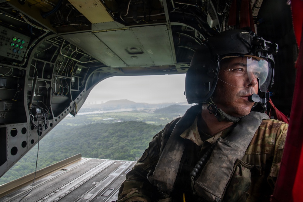 A military aircrew member is seated inside what appears to be a large military helicopter, likely a CH-47 Chinook, gazing out through the open rear ramp area. The individual is wearing full flight gear including a flight helmet with a visor, communications microphone, and a tactical vest over a camouflage uniform. Below and beyond the aircraft, a lush green landscape with hills and what appears to be a coastal or river area is visible.