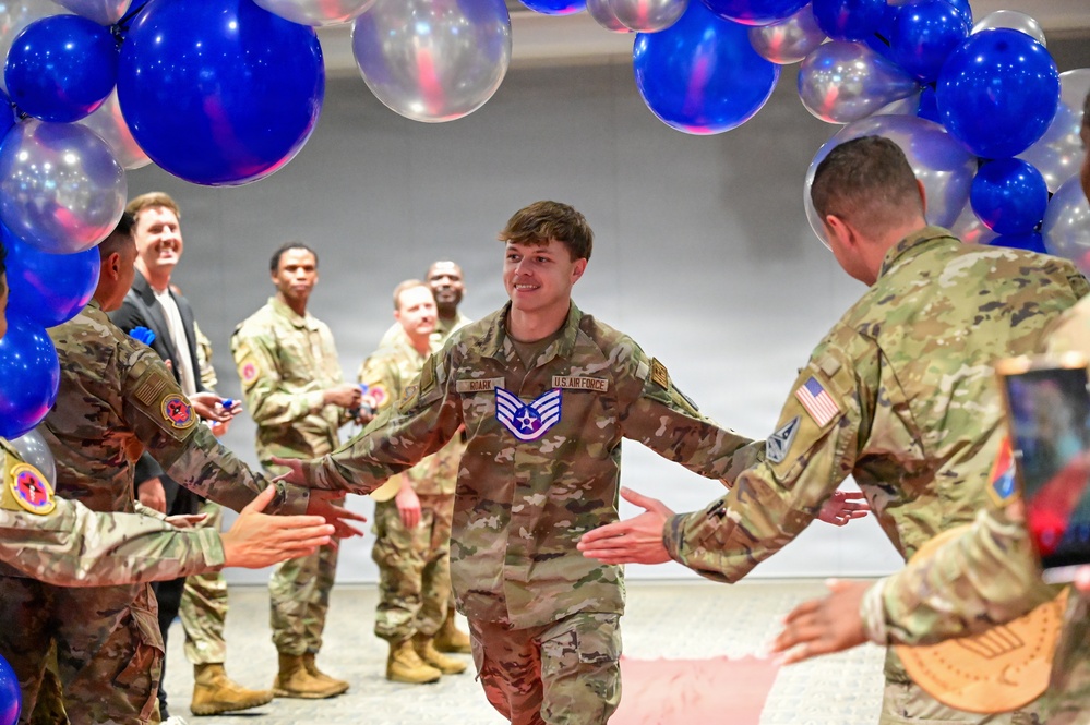 A U.S. Air Force Staff Sergeant walks through a congratulatory gauntlet formed by fellow service members under a decorative arch of blue, red, and clear balloons. The airman is smiling and exchanging high-fives with colleagues on both sides, suggesting a recognition or promotion ceremony. The setting appears to be an indoor military event space decorated for a celebratory occasion.