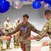 A U.S. Air Force Staff Sergeant walks through a congratulatory gauntlet formed by fellow service members under a decorative arch of blue, red, and clear balloons. The airman is smiling and exchanging high-fives with colleagues on both sides, suggesting a recognition or promotion ceremony. The setting appears to be an indoor military event space decorated for a celebratory occasion.