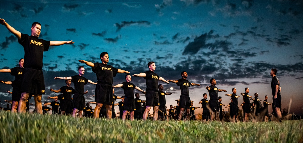 A large group of U.S. Army soldiers in black PT (Physical Training) uniforms are performing synchronized exercises on a grass field at dusk or dawn. The soldiers are arranged in rows extending into the background, all with arms outstretched laterally in a coordinated drill position. The dramatic twilight sky with scattered clouds provides a striking backdrop to the formation.