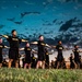 A large group of U.S. Army soldiers in black PT (Physical Training) uniforms are performing synchronized exercises on a grass field at dusk or dawn. The soldiers are arranged in rows extending into the background, all with arms outstretched laterally in a coordinated drill position. The dramatic twilight sky with scattered clouds provides a striking backdrop to the formation.