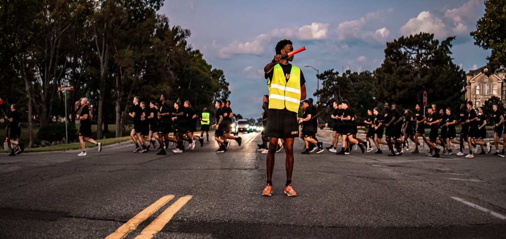 A large group of individuals dressed in matching black athletic uniforms are running in formation along a road at dusk or early morning. A person wearing a high-visibility yellow safety vest stands in the center of the road holding a red baton or light wand, appearing to serve as a safety marshal or guide. The scene strongly suggests a military physical training run, with 'ARMY' visible on the central figure's shorts.