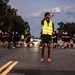 A large group of individuals dressed in matching black athletic uniforms are running in formation along a road at dusk or early morning. A person wearing a high-visibility yellow safety vest stands in the center of the road holding a red baton or light wand, appearing to serve as a safety marshal or guide. The scene strongly suggests a military physical training run, with 'ARMY' visible on the central figure's shorts.