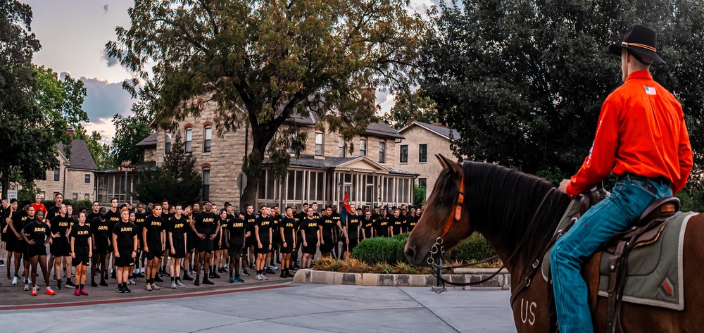 A large formation of Army personnel wearing black 'ARMY' PT (physical training) shirts and shorts stand at attention in an outdoor plaza area. A rider on horseback, wearing a bright orange shirt and cowboy hat, faces the formation from the right side of the frame. The setting appears to be a historic military installation with stone buildings visible in the background, likely during early morning or evening hours.