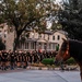 A large formation of Army personnel wearing black 'ARMY' PT (physical training) shirts and shorts stand at attention in an outdoor plaza area. A rider on horseback, wearing a bright orange shirt and cowboy hat, faces the formation from the right side of the frame. The setting appears to be a historic military installation with stone buildings visible in the background, likely during early morning or evening hours.