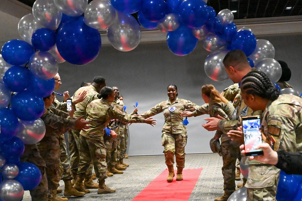 A military service member in camouflage uniform walks down a red carpet through a celebratory gauntlet of fellow service members under a decorative arch of blue, red, and silver balloons. The central figure appears to be the honoree of a promotion or recognition ceremony, smiling broadly with arms outstretched as colleagues reach out to congratulate her. Bystanders are capturing the moment on smartphones.