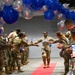 A military service member in camouflage uniform walks down a red carpet through a celebratory gauntlet of fellow service members under a decorative arch of blue, red, and silver balloons. The central figure appears to be the honoree of a promotion or recognition ceremony, smiling broadly with arms outstretched as colleagues reach out to congratulate her. Bystanders are capturing the moment on smartphones.