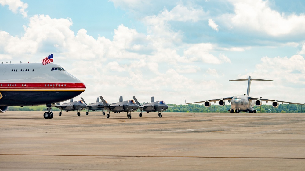 The image shows a military flight line featuring multiple aircraft parked on a tarmac. A large wide-body aircraft with a distinctive red, white, and blue livery and an American flag is visible on the left, flanked by three F-35 stealth fighter jets in the center, and a large military transport aircraft (C-17 Globemaster III) on the right. The scene appears to be at a U.S. military airfield under partly cloudy skies.