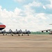 The image shows a military flight line featuring multiple aircraft parked on a tarmac. A large wide-body aircraft with a distinctive red, white, and blue livery and an American flag is visible on the left, flanked by three F-35 stealth fighter jets in the center, and a large military transport aircraft (C-17 Globemaster III) on the right. The scene appears to be at a U.S. military airfield under partly cloudy skies.