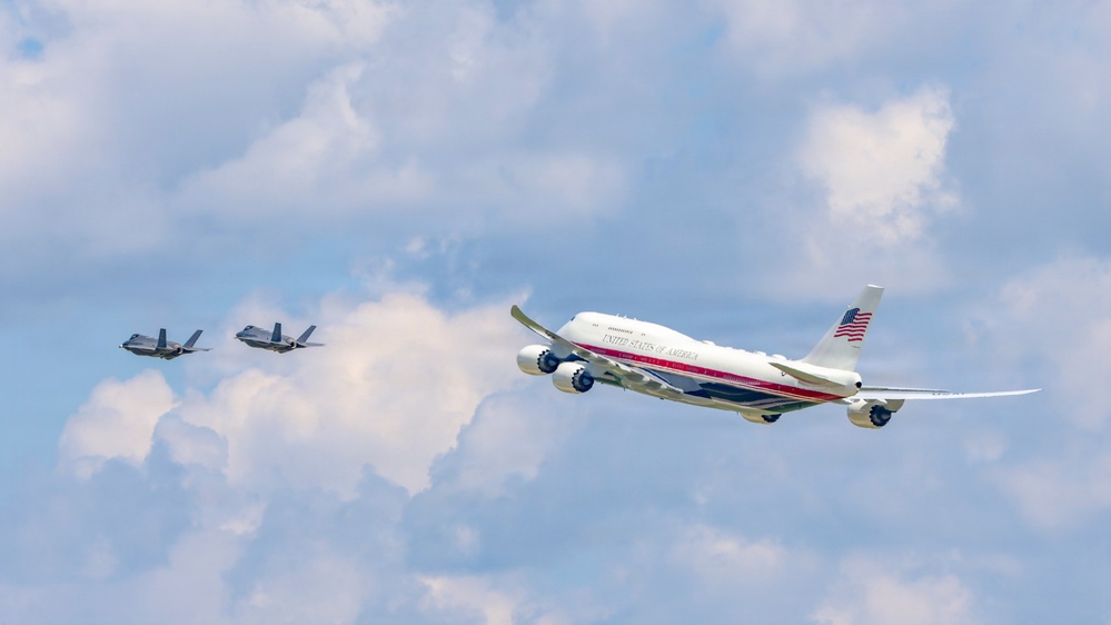 A large white aircraft marked 'United States of America' with red and gold striping and an American flag on the tail flies in formation with two smaller military fighter jets against a partly cloudy sky. The large aircraft appears to be a Boeing 747-style wide-body jet in a presidential or government livery. Two fighter jets, consistent with F-35 or similar stealth aircraft, flank the larger plane to its left rear.