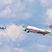 A large white aircraft marked 'United States of America' with red and gold striping and an American flag on the tail flies in formation with two smaller military fighter jets against a partly cloudy sky. The large aircraft appears to be a Boeing 747-style wide-body jet in a presidential or government livery. Two fighter jets, consistent with F-35 or similar stealth aircraft, flank the larger plane to its left rear.