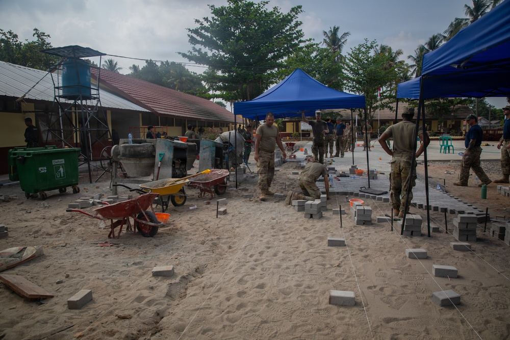 A construction or civic action project is underway in what appears to be a tropical compound or school courtyard. Multiple workers, some in military-style uniforms, are laying concrete paving blocks on a sand base. The site includes equipment such as wheelbarrows, a cement mixer, and blue canopy tents providing shade.