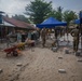 A construction or civic action project is underway in what appears to be a tropical compound or school courtyard. Multiple workers, some in military-style uniforms, are laying concrete paving blocks on a sand base. The site includes equipment such as wheelbarrows, a cement mixer, and blue canopy tents providing shade.