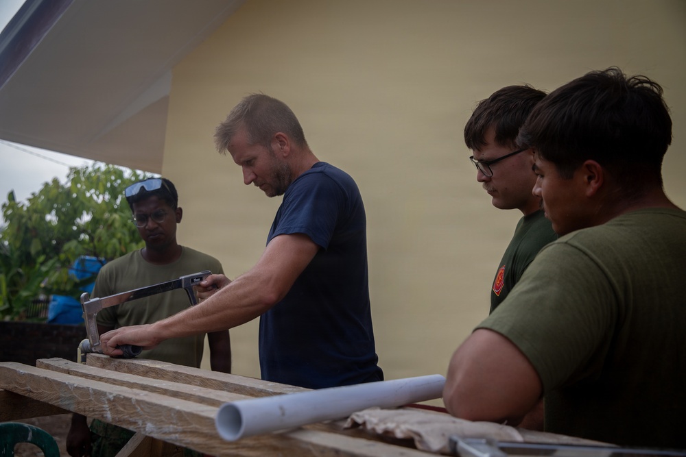 A man in a navy blue t-shirt is demonstrating how to use a hacksaw on a wooden plank to three other individuals wearing olive/military-green t-shirts. The group appears to be engaged in a construction or carpentry task outdoors, near a yellow-painted building. A white PVC pipe is visible on the work surface, suggesting plumbing or structural work is underway.