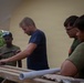 A man in a navy blue t-shirt is demonstrating how to use a hacksaw on a wooden plank to three other individuals wearing olive/military-green t-shirts. The group appears to be engaged in a construction or carpentry task outdoors, near a yellow-painted building. A white PVC pipe is visible on the work surface, suggesting plumbing or structural work is underway.