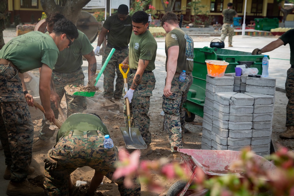 A group of military personnel in camouflage uniforms are engaged in construction or cleanup work outdoors. They are using shovels and other tools to move dirt and debris, with a stack of concrete blocks and a green dumpster visible nearby. The setting appears to be a humanitarian assistance or civic action project at a compound or base.