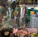 A group of military personnel in camouflage uniforms are engaged in construction or cleanup work outdoors. They are using shovels and other tools to move dirt and debris, with a stack of concrete blocks and a green dumpster visible nearby. The setting appears to be a humanitarian assistance or civic action project at a compound or base.