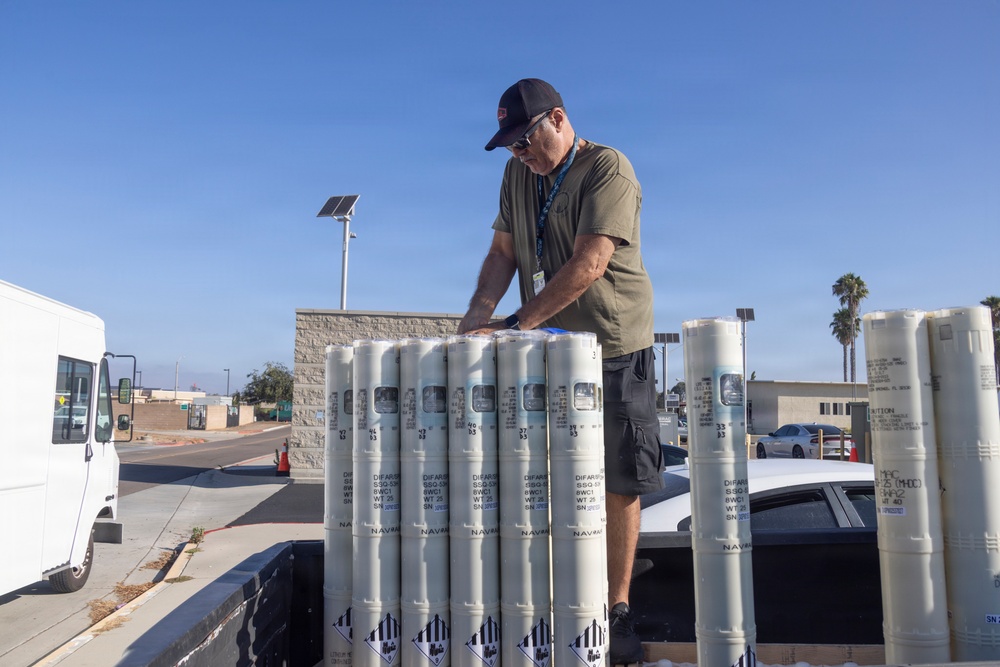 A man wearing a black cap, sunglasses, olive t-shirt, and an ID lanyard is handling a group of cylindrical white military/naval ordnance containers on what appears to be a loading dock or flatbed. The containers are labeled with 'NAVR' markings and display hazardous materials (explosive) warning diamonds at their bases. The setting appears to be a military or government facility in a sunny, arid environment with palm trees visible in the background.