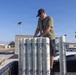 A man wearing a black cap, sunglasses, olive t-shirt, and an ID lanyard is handling a group of cylindrical white military/naval ordnance containers on what appears to be a loading dock or flatbed. The containers are labeled with 'NAVR' markings and display hazardous materials (explosive) warning diamonds at their bases. The setting appears to be a military or government facility in a sunny, arid environment with palm trees visible in the background.
