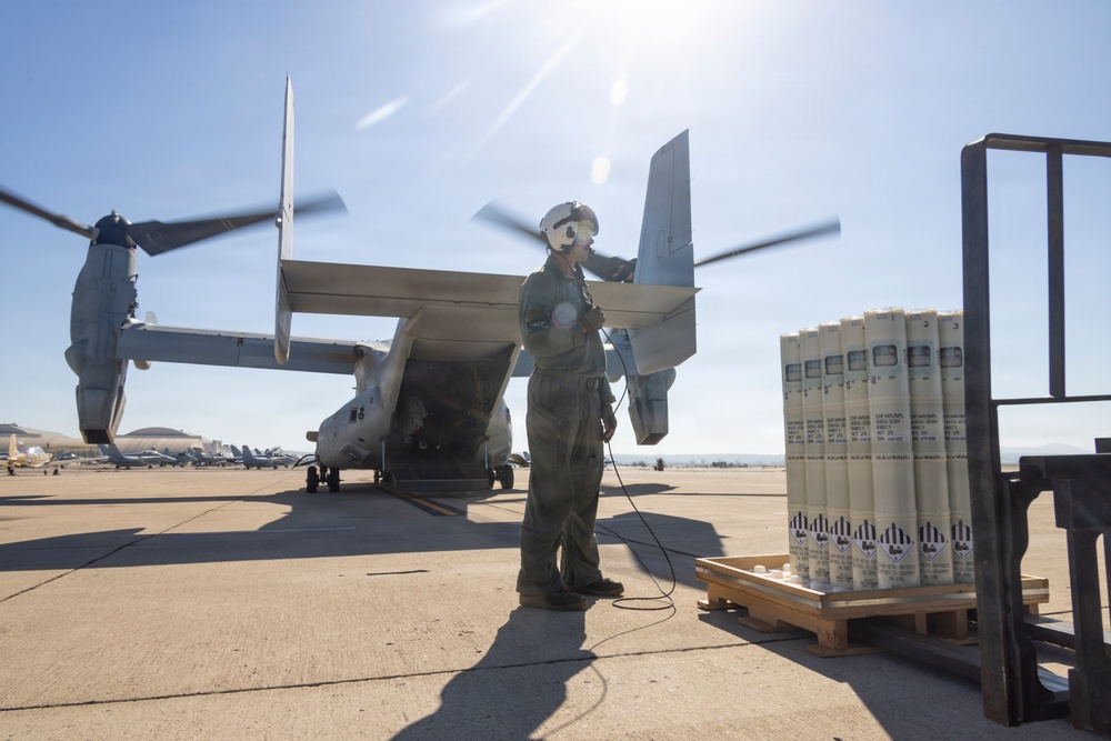 A military aircrew member in flight suit and helmet stands on a tarmac next to a V-22 Osprey tiltrotor aircraft with rotors spinning. A pallet of cylindrical munitions or ordnance containers is staged to the right of the personnel. The scene appears to be at a military air base under bright, clear sky conditions.