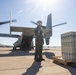 A military aircrew member in flight suit and helmet stands on a tarmac next to a V-22 Osprey tiltrotor aircraft with rotors spinning. A pallet of cylindrical munitions or ordnance containers is staged to the right of the personnel. The scene appears to be at a military air base under bright, clear sky conditions.