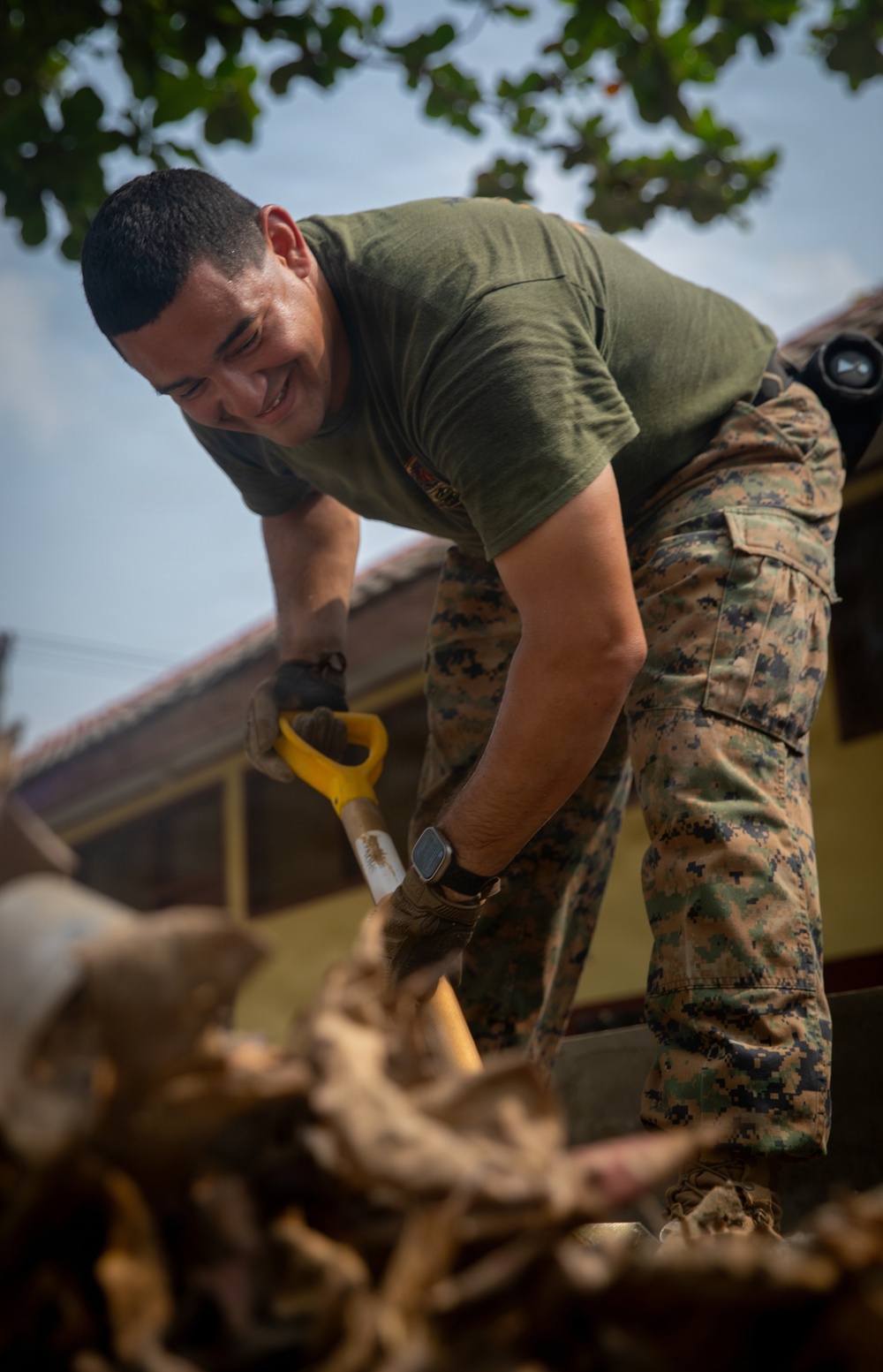 A smiling military service member wearing MARPAT camouflage trousers and an olive green t-shirt is actively working with a hand saw, cutting through what appears to be wood or debris. The individual appears to be engaged in a construction, cleanup, or humanitarian assistance task in an outdoor setting near a building. The mood is positive, as indicated by the subject's visible smile.