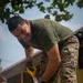 A smiling military service member wearing MARPAT camouflage trousers and an olive green t-shirt is actively working with a hand saw, cutting through what appears to be wood or debris. The individual appears to be engaged in a construction, cleanup, or humanitarian assistance task in an outdoor setting near a building. The mood is positive, as indicated by the subject's visible smile.
