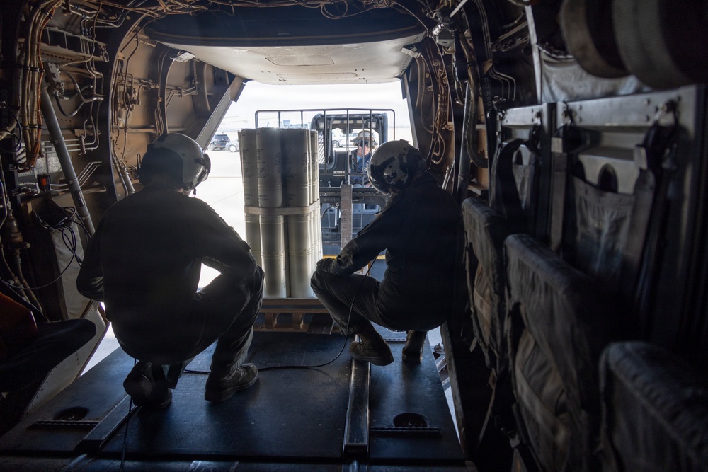 Two personnel wearing flight helmets are crouched inside what appears to be a military cargo aircraft, observing cylindrical cargo being loaded via forklift through the rear ramp opening. A third individual is visible operating the forklift outside the aircraft. The interior structural elements of the cargo bay are clearly visible, including wiring, tie-down rails, and padded sidewalls.