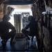 Two personnel wearing flight helmets are crouched inside what appears to be a military cargo aircraft, observing cylindrical cargo being loaded via forklift through the rear ramp opening. A third individual is visible operating the forklift outside the aircraft. The interior structural elements of the cargo bay are clearly visible, including wiring, tie-down rails, and padded sidewalls.