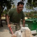 A male service member wearing U.S. Marine Corps MARPAT camouflage trousers and an olive green t-shirt is loading or moving broken concrete blocks using a red wheelbarrow. He is wearing safety glasses and has visible tattoos on both arms. The setting appears to be an outdoor construction or cleanup site in a tropical environment.