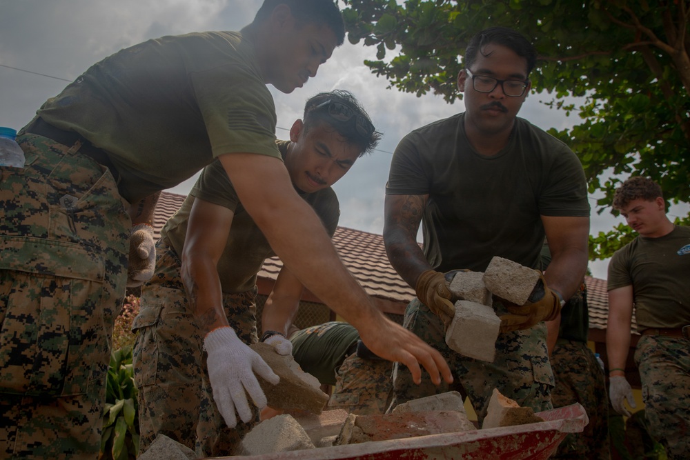Military personnel in camouflage uniforms are engaged in construction or debris-clearing work, handling concrete blocks and rubble. The scene appears to be a humanitarian assistance or engineering project near a building with a tiled roof. At least four service members are visible, working collaboratively to move or sort broken masonry materials.