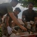 Military personnel in camouflage uniforms are engaged in construction or debris-clearing work, handling concrete blocks and rubble. The scene appears to be a humanitarian assistance or engineering project near a building with a tiled roof. At least four service members are visible, working collaboratively to move or sort broken masonry materials.
